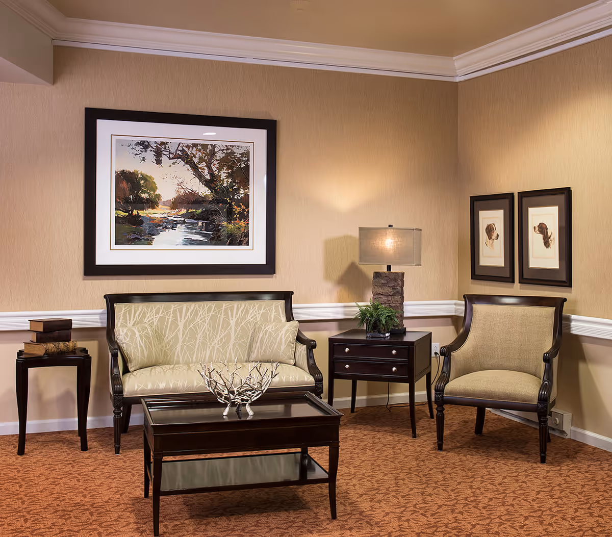 A cozy sitting area with a beige patterned loveseat and matching armchair, a dark wooden coffee table with a decorative centerpiece, a side table with stacked books, and a dark wooden nightstand with a lamp and a small plant. The walls are beige with white trim and decorated with framed artwork, including a large landscape painting and two smaller portraits.