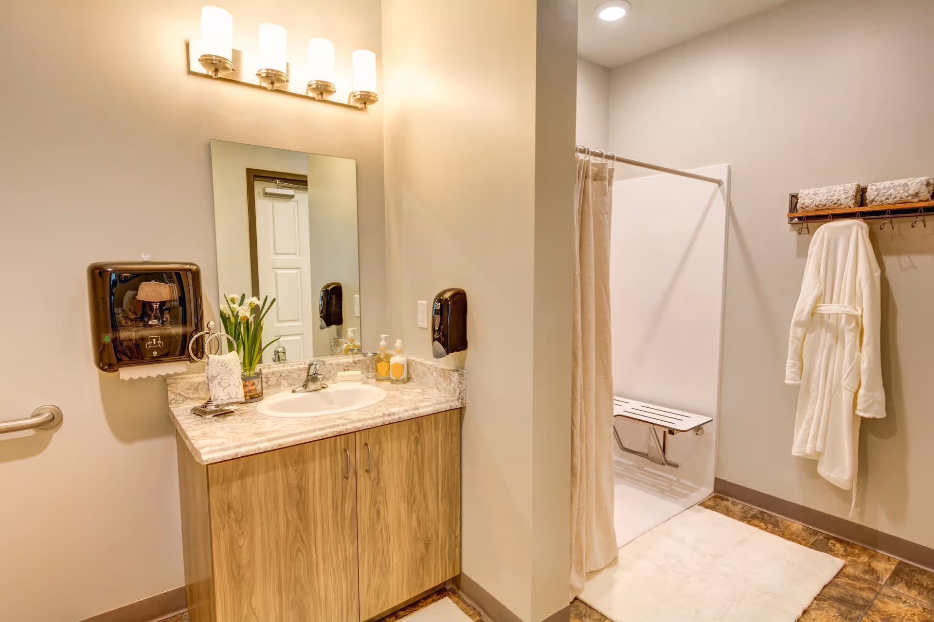 A clean and well-lit bathroom featuring a wooden vanity with a marble countertop, a sink, a mirror above the sink, and a wall-mounted light fixture with four lights. To the right, there is a walk-in shower with a fold-down seat and a beige shower curtain. A white bathrobe hangs on a wall-mounted rack with towels above it. The floor has a brown tile pattern and a white bath mat is placed in front of the shower.