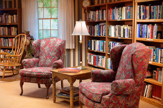 Cozy library seating area with two patterned wingback chairs, a side table with a lamp, and bookshelves by a window.