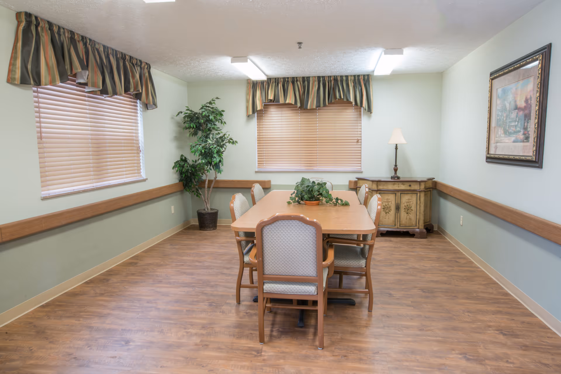 A small dining room with a rectangular wooden table surrounded by six chairs. The room has light green walls with wooden trim, two windows with closed blinds and striped valances, a potted plant in the corner, a wooden sideboard with a lamp, and a framed painting on the wall.