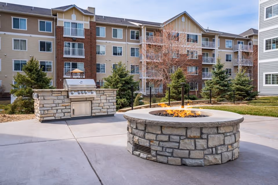 Outdoor patio area at Hayden Grove Bloomington featuring a stone fire pit with flames and a built-in stainless steel grill. The background shows a multi-story residential building with balconies and windows, surrounded by trees and grass.