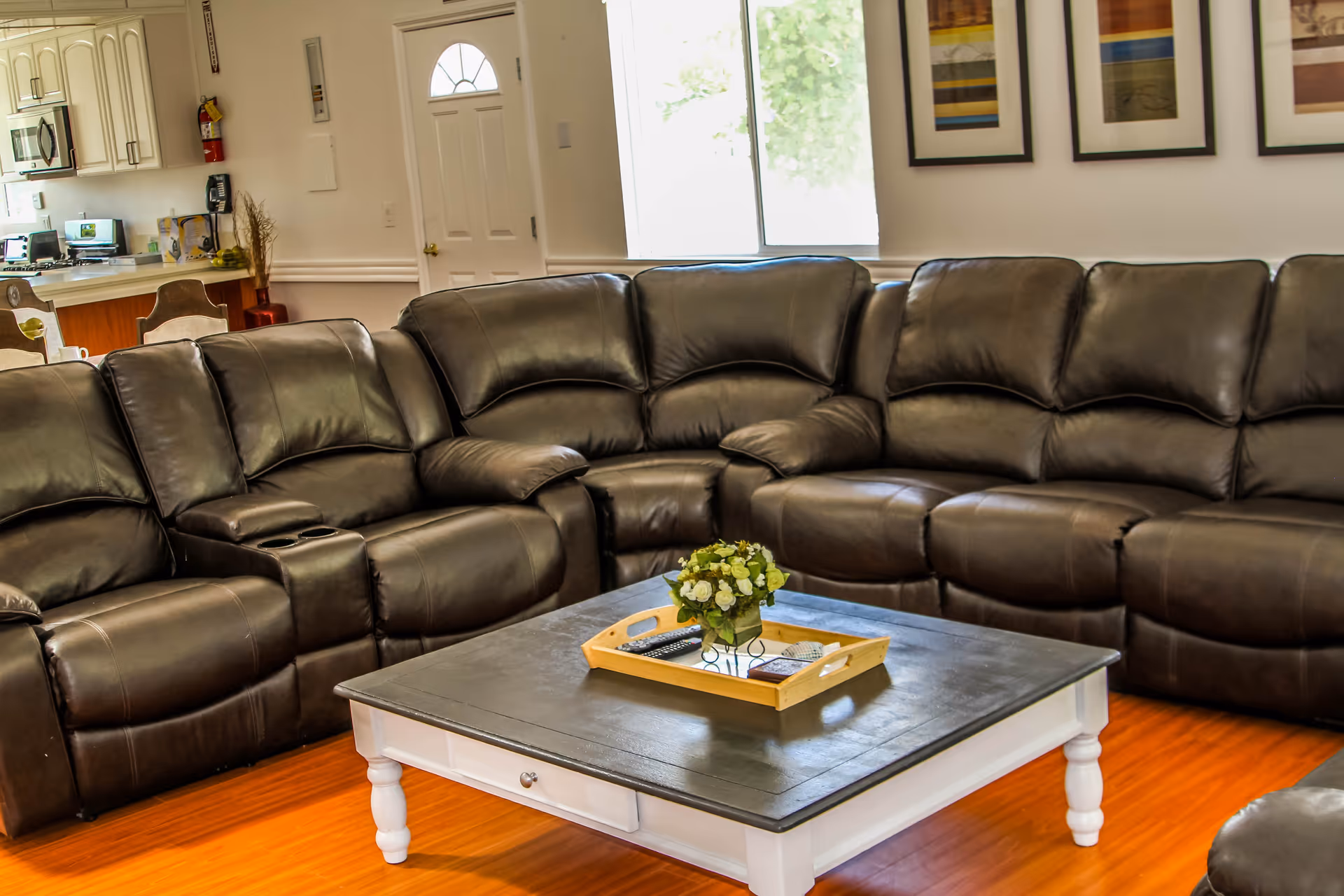 A cozy living room with a large dark brown leather sectional sofa arranged around a square coffee table with a black top and white legs. On the coffee table is a wooden tray holding a small green plant, a remote control, and some other small items. The room has wooden flooring, a window letting in natural light, and framed artwork on the wall. In the background, a kitchen area with white cabinets and appliances is partially visible.