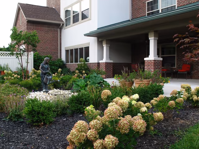A garden area with blooming hydrangea bushes and other greenery in front of a brick and white building. There is a statue of a girl holding a watering can among the plants. The building has a covered porch with red chairs and large windows.