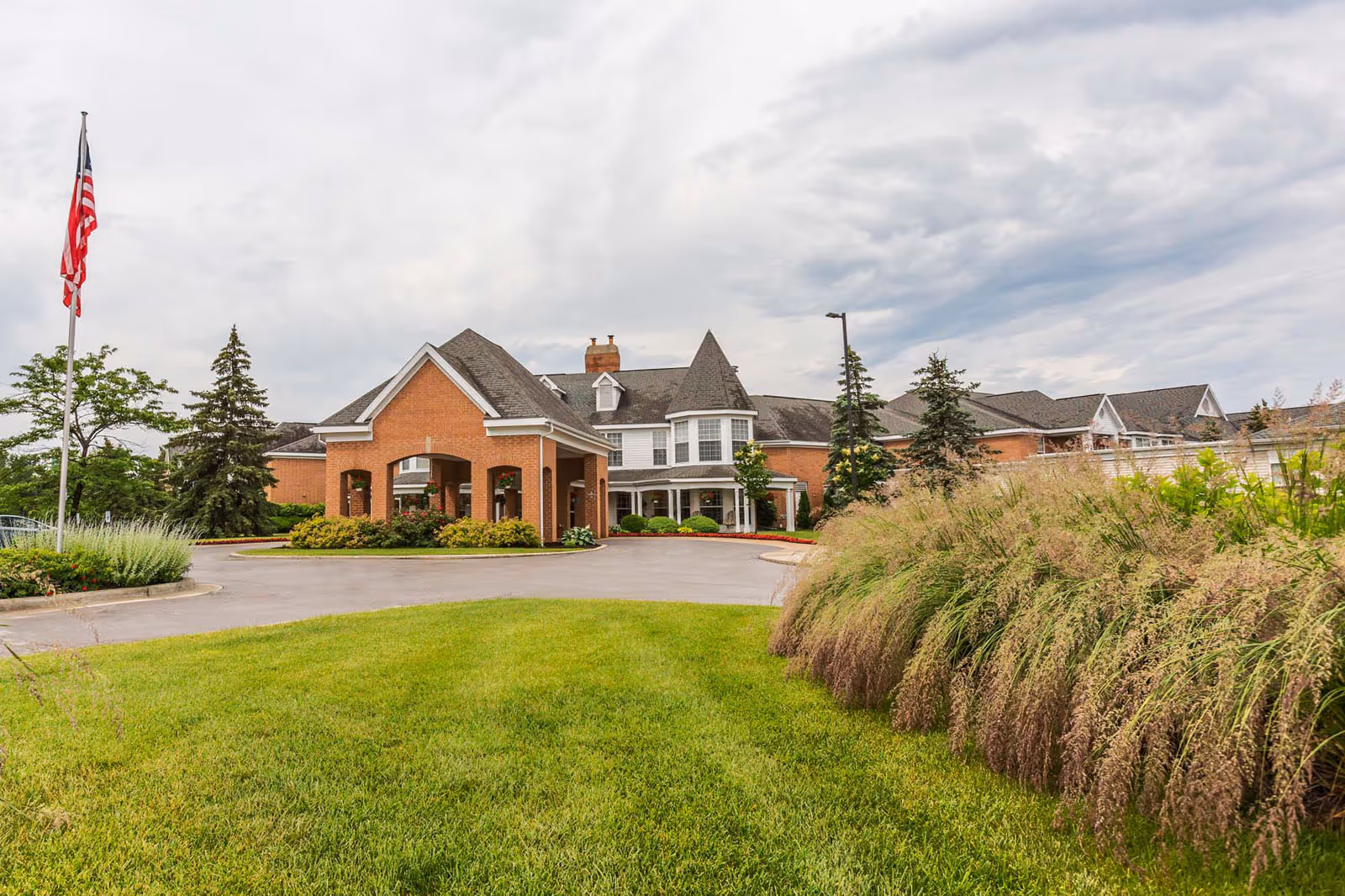 Front exterior of a brick senior living building with a covered entrance, driveway, flagpole, and landscaped lawn under a cloudy sky.