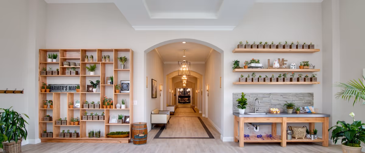 A bright and spacious interior hallway with light-colored walls and wood flooring. On the left side, there is a large wooden shelving unit filled with small potted plants and decorative items, including a sign that reads 'FARMER'S MARKET.' On the right side, there are two wooden shelves mounted on the wall holding more small potted plants and decorative items, with a wooden countertop below featuring a sink and additional plants. The hallway extends into the distance with modern light fixtures hanging from the ceiling and seating areas along the walls.