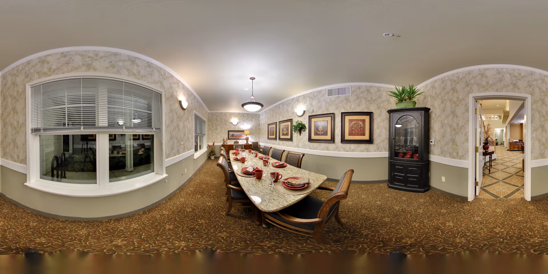 A dining room with a long rectangular table set with red plates, bowls, and cups. The room has patterned carpet, wallpapered walls with framed artwork, a black display cabinet with plants on top, and a large window with blinds. There is a doorway leading to another room with tiled flooring.