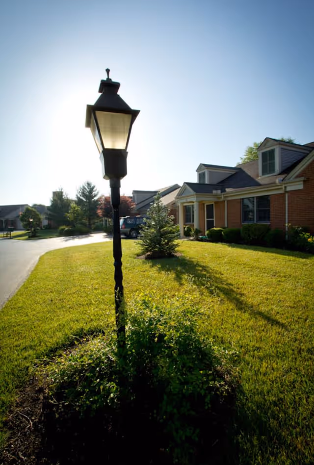 A black vintage-style street lamp stands in a small garden bed surrounded by green grass in front of a brick building with white trim and dormer windows under a clear blue sky.