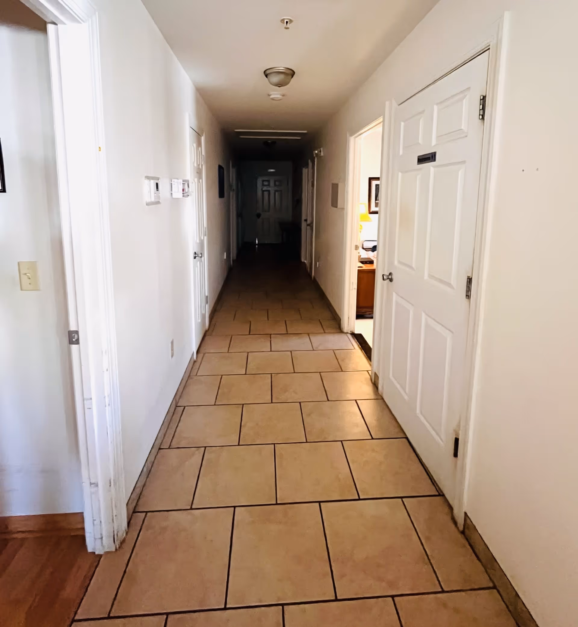 A long hallway with tiled flooring and white walls, featuring several closed doors on either side, leading to a distant entrance.