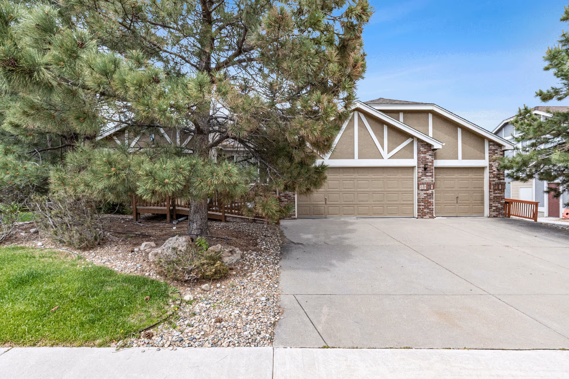 Exterior view of a residential building with a three-car garage, surrounded by trees and landscaping with rocks and grass under a clear blue sky.