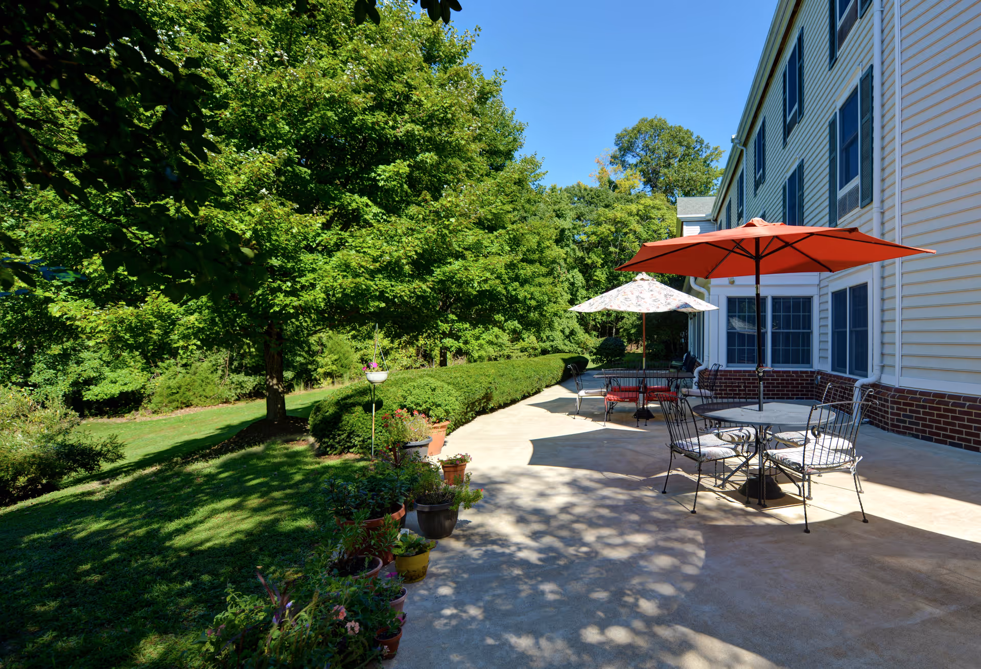 Outdoor patio area at Charter Senior Living of Fredericksburg with tables and chairs under large umbrellas, surrounded by greenery and trees on a sunny day.