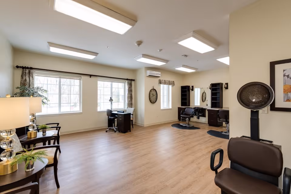 A spacious, well-lit room with large windows, wooden flooring, and beige walls. The room contains salon chairs with mirrors and hairdressing stations, a hair dryer chair, a small desk with a chair near the window, and a seating area with two chairs and a side table with lamps and plants.