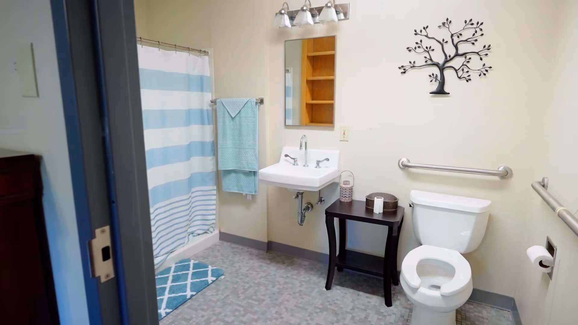 A bathroom with a white toilet, a white sink with two handles, a mirror above the sink, a small dark wooden table holding a roll of toilet paper and a decorative item, a blue and white striped shower curtain, a blue bath mat, and a blue towel hanging on a towel rack. There are grab bars on the walls near the toilet, and a decorative metal tree wall art above the toilet.