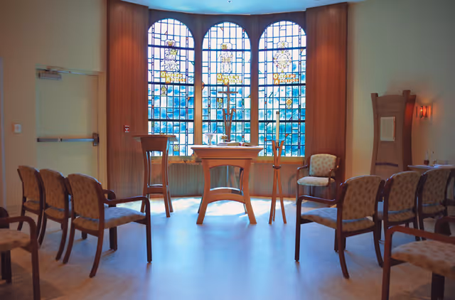 A small chapel or meditation room with wooden chairs arranged in rows facing a wooden altar table. Behind the altar are three tall stained glass windows with religious imagery and text. The room has soft lighting and a calm atmosphere.