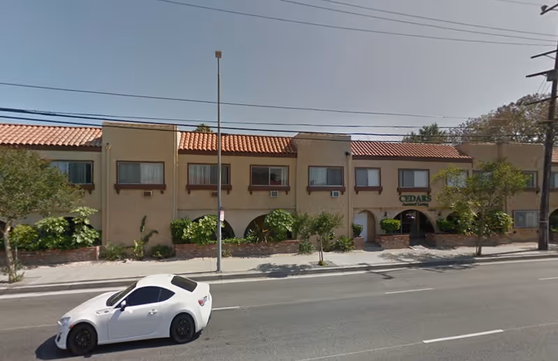 Exterior view of Cedars Assisted Living facility, a two-story building with beige stucco walls and a red tile roof. There are several windows with air conditioning units and some greenery in front of the building. A white car is parked on the street in front of the facility.