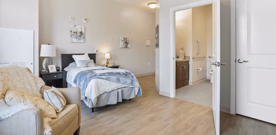 A cozy bedroom in a senior living facility with a single bed covered in white and blue bedding, a nightstand with a lamp and picture frame, a beige armchair with pillows and a throw blanket, and an open door leading to a bathroom with a toilet and sink visible.