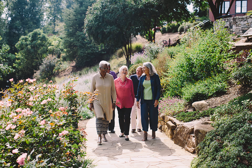 A group of four elderly people walking and chatting along a stone pathway surrounded by lush greenery and blooming flowers in an outdoor garden setting.