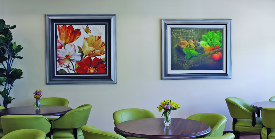 Bright common dining area with round wooden tables, green chairs, small vases of flowers, and framed artwork on a pale wall.