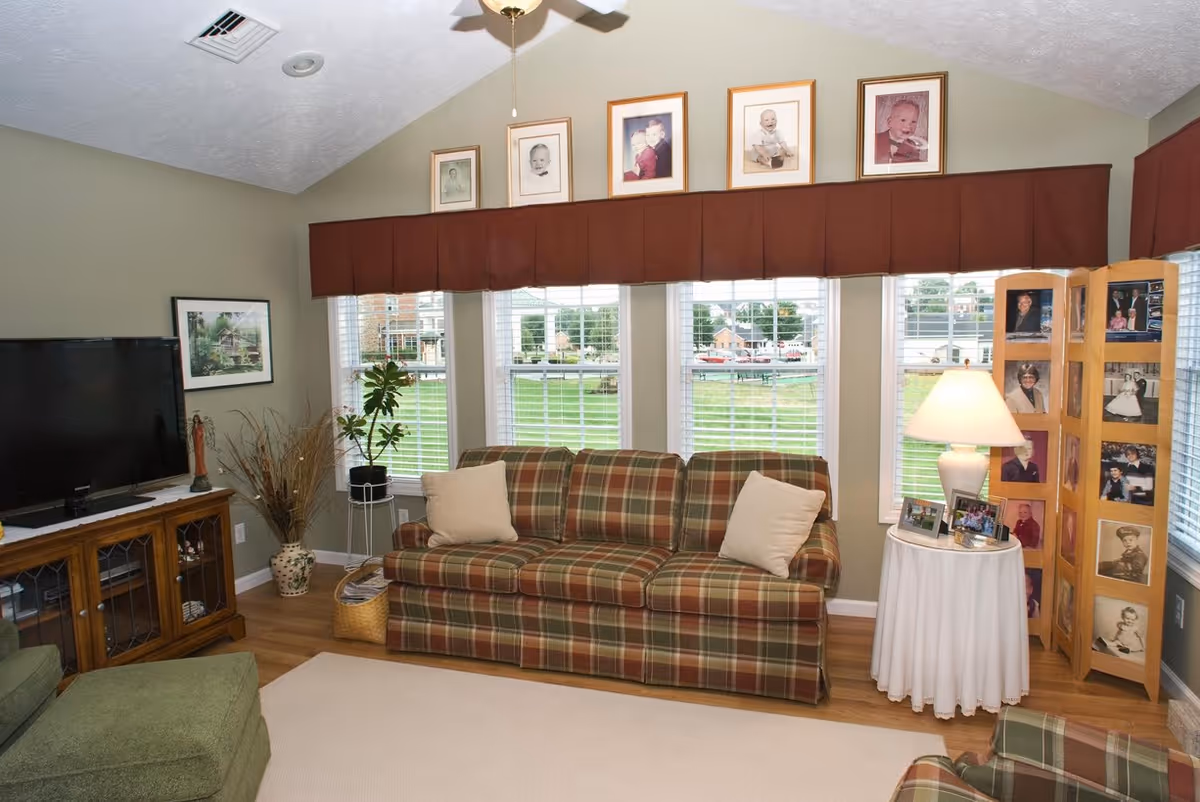 A cozy living room with a plaid sofa and two beige pillows, a green armchair, a wooden TV stand with a flat-screen TV, a small round table with a white tablecloth and a lamp, and a wooden photo display with framed pictures. The room has large windows with white blinds and a brown valance, and several framed baby photos are displayed above the windows.