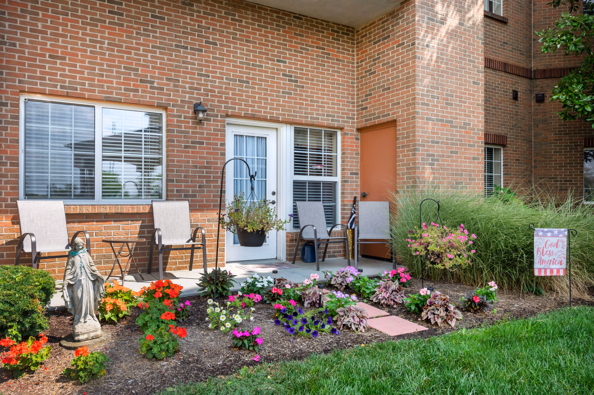 Outdoor patio area of a brick building with four chairs, a small table, hanging flower pots, and a garden bed with colorful flowers and a statue. A sign in the garden reads 'God Bless America.'