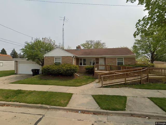 Single-story brick house with a brown roof, a front porch with a wooden wheelchair ramp, green bushes, and trees on a grassy lawn. There is a concrete sidewalk and driveway leading to a detached garage on the left side.