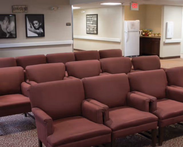 Rows of maroon upholstered chairs arranged theater-style in a communal room with framed wall art and a small kitchenette area in the background.
