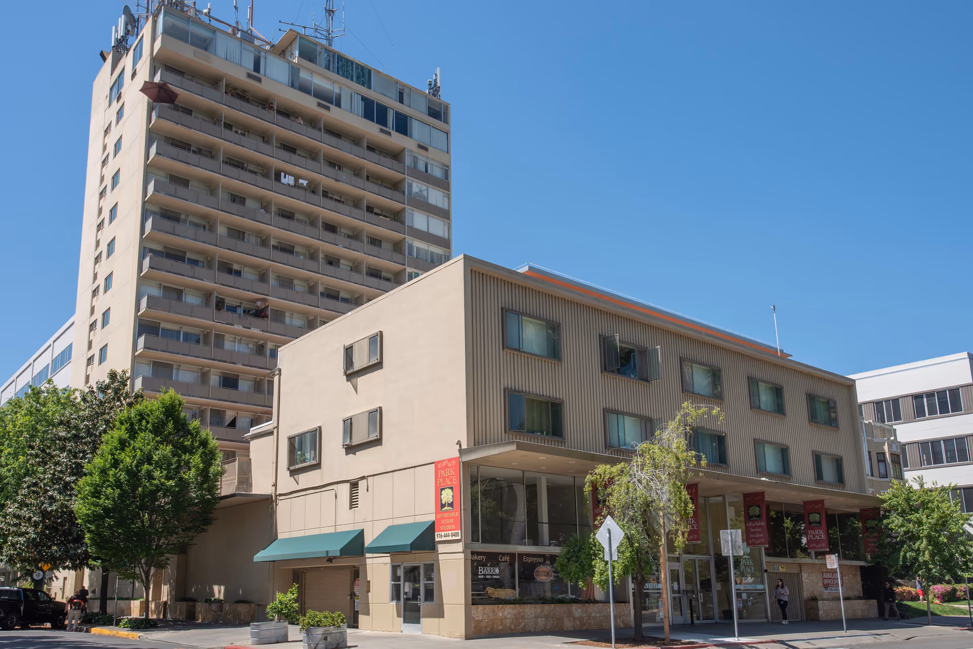 Exterior view of Park Place Apartments, a multi-story beige building with balconies and several windows, under a clear blue sky. The building has red signs indicating affordable senior studios and a cafe on the ground floor. Trees and a few people are visible near the entrance.