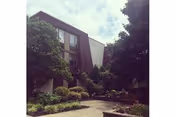 Three-story building with a recessed entrance, landscaped shrubs and trees, and a paved walkway in front.