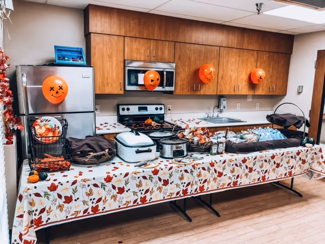 A kitchen area decorated for Halloween with orange balloons featuring jack-o'-lantern faces attached to the cabinets and refrigerator. A long table covered with a fall-themed tablecloth holds various food items, including covered dishes, condiments, and bottled water. The kitchen has wooden cabinets, a stainless steel refrigerator, a microwave, and a stove.