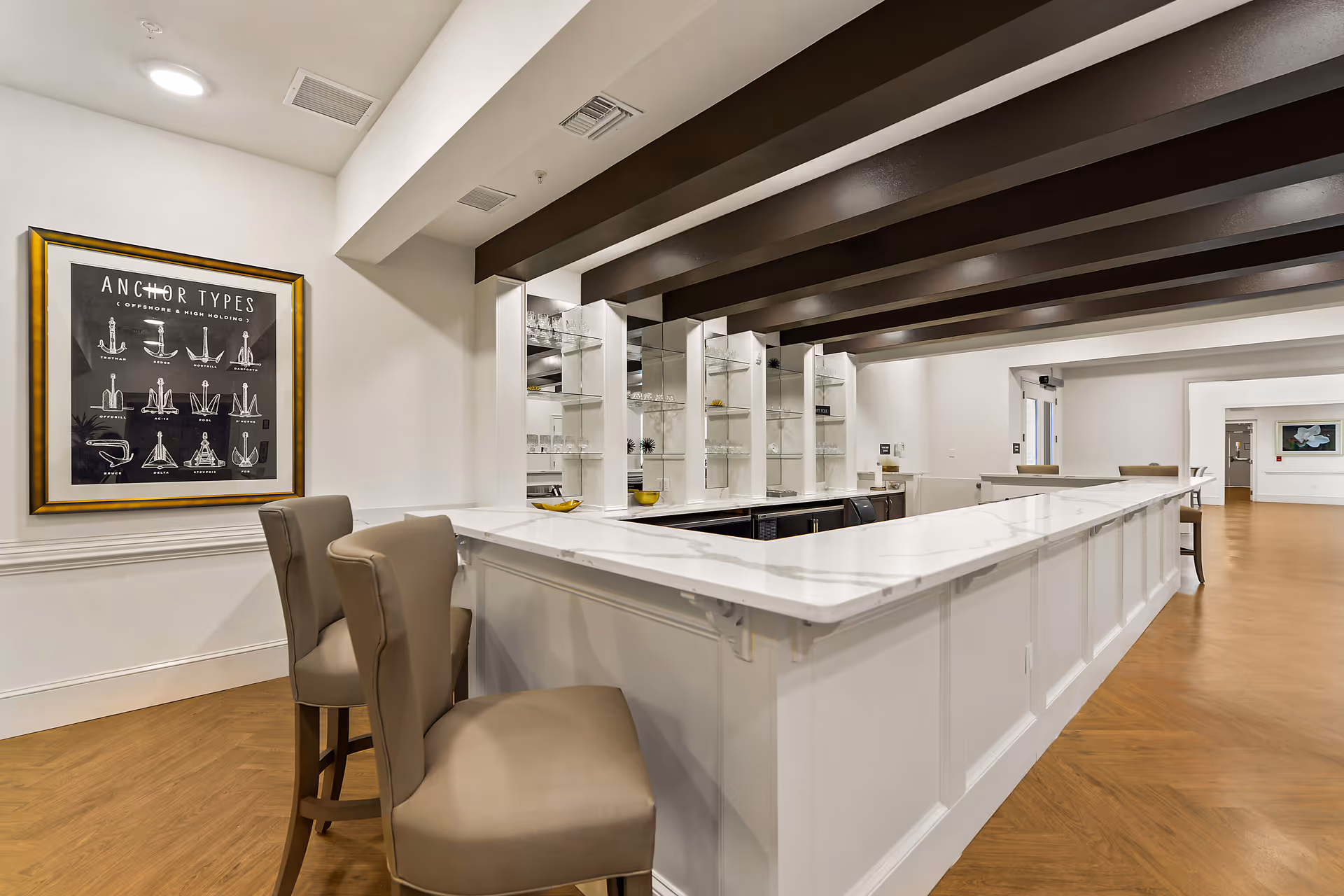 Interior view of a modern bar area with a white marble countertop, beige cushioned bar stools, and glass shelves with glassware. The ceiling has dark wooden beams, and there is a framed poster on the wall titled 'Anchor Types'. The floor is wooden, and the space extends into a larger room with additional seating.