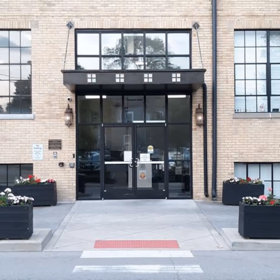 Front entrance of a brick building with glass double doors, large windows, planter boxes, and a crosswalk leading to the doorway.