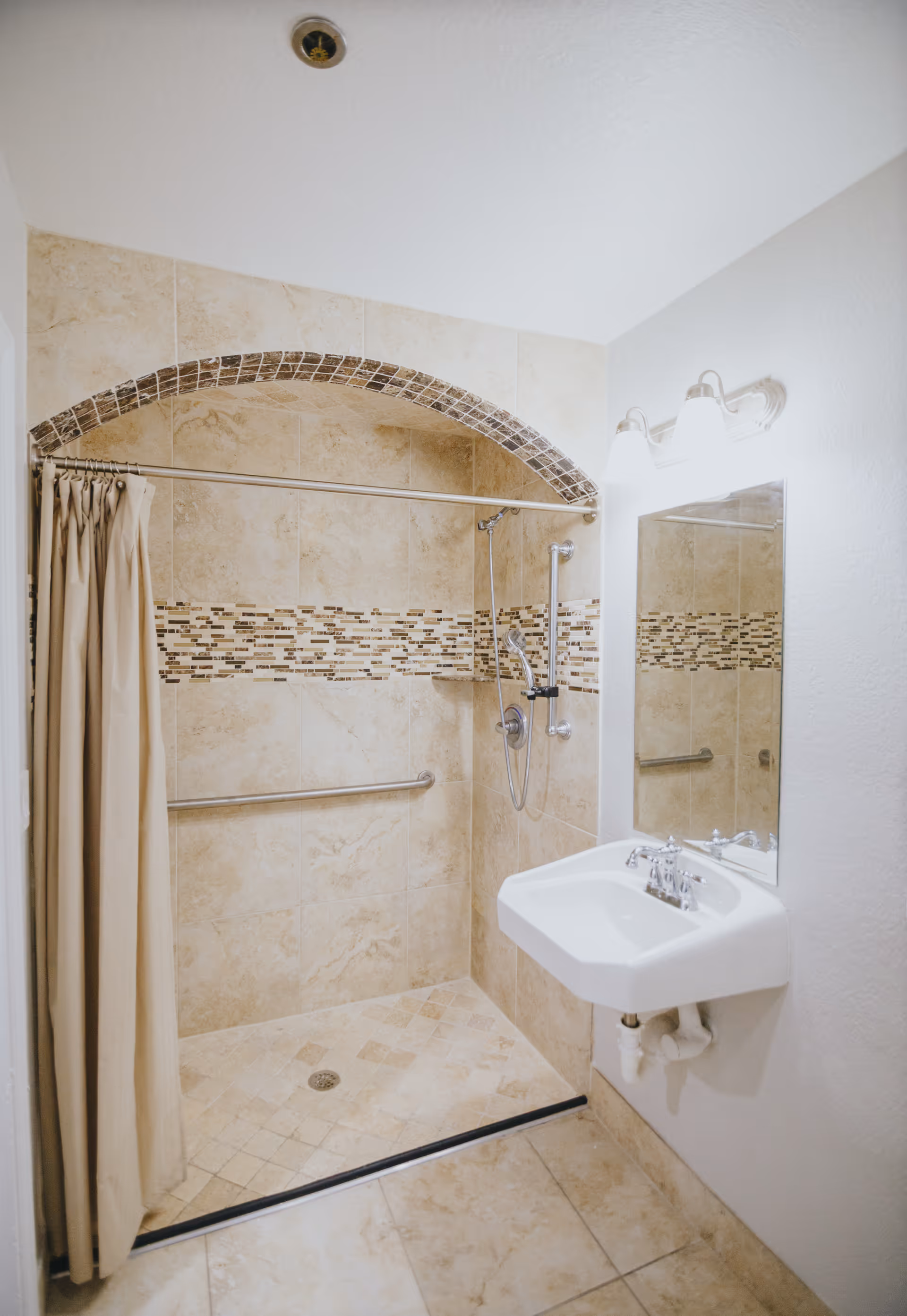 Accessible tiled bathroom with a walk-in shower featuring a curtain, grab bars, handheld showerhead, and a wall-mounted sink with mirror and light fixture.