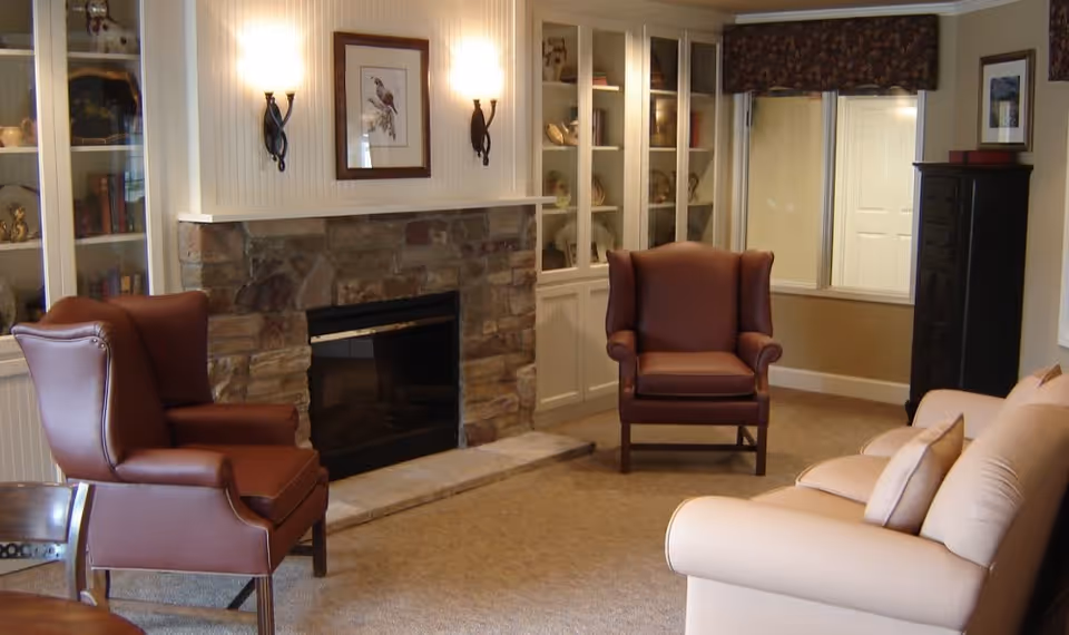 A cozy living room with a stone fireplace in the center, flanked by built-in white cabinets with shelves displaying decorative items. There are two brown leather armchairs facing the fireplace and a beige sofa with cushions on the right side. The room has beige carpet, wall sconces lighting the fireplace area, and a window with a valance on the far wall.