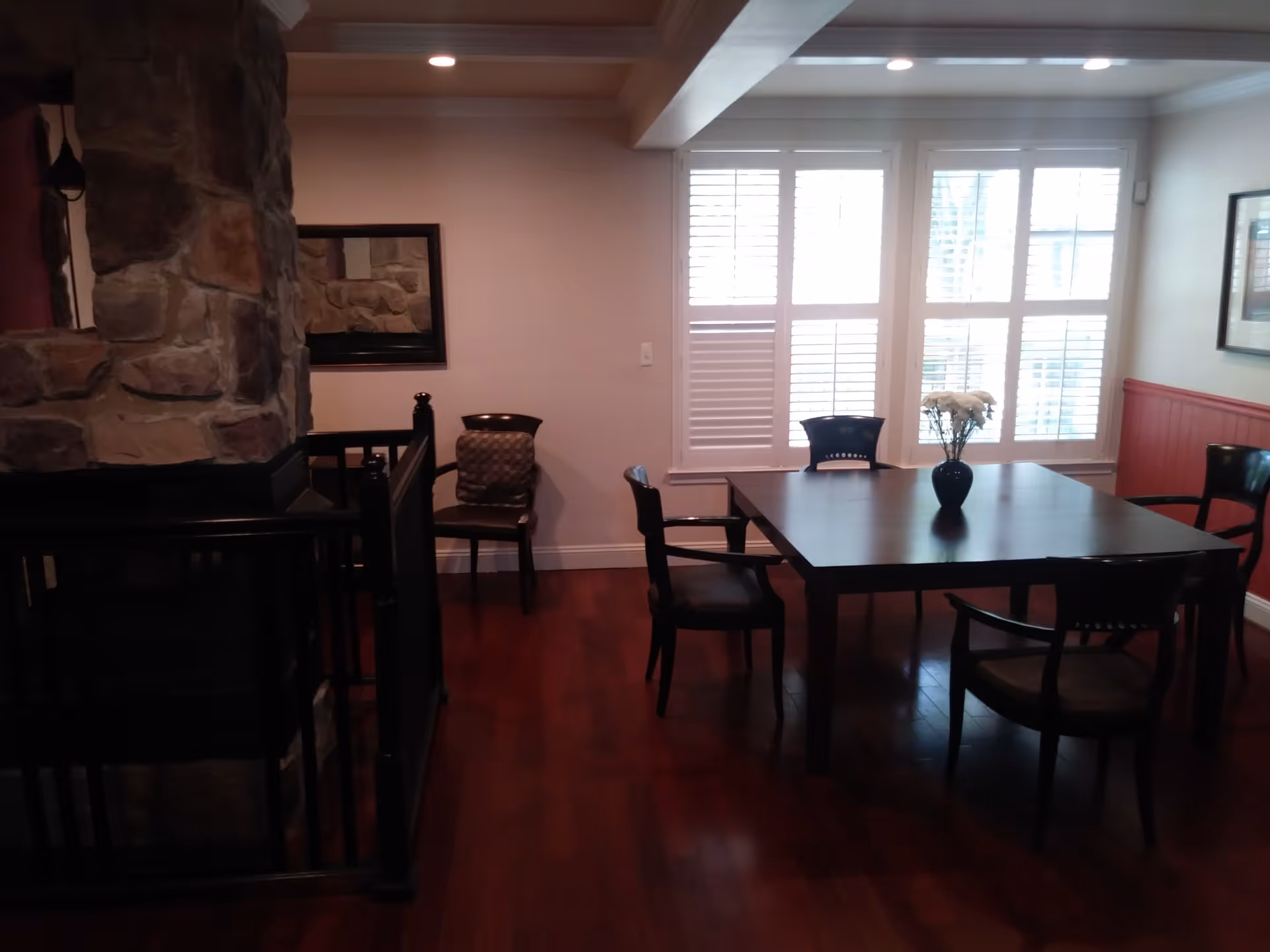 Interior view of a dining area with a dark wooden table surrounded by five chairs. A vase with white flowers is placed on the table. The room has wooden flooring, a stone fireplace on the left, and large windows with white shutters letting in natural light. A framed picture hangs on the right wall.