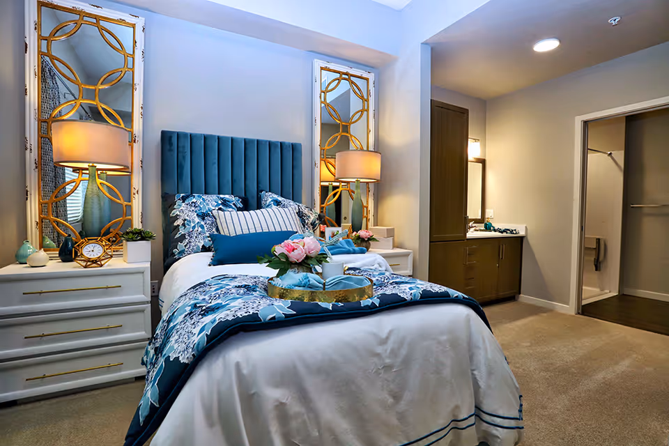 A cozy bedroom in a senior living facility featuring a bed with a blue upholstered headboard, floral bedding, and decorative pillows. On either side of the bed are white nightstands with gold handles, each topped with a lamp and decorative items. Behind the nightstands are tall mirrors with gold geometric frames. To the right, there is a vanity area with a sink and mirror, and an open doorway leading to a bathroom with a shower and grab bars.