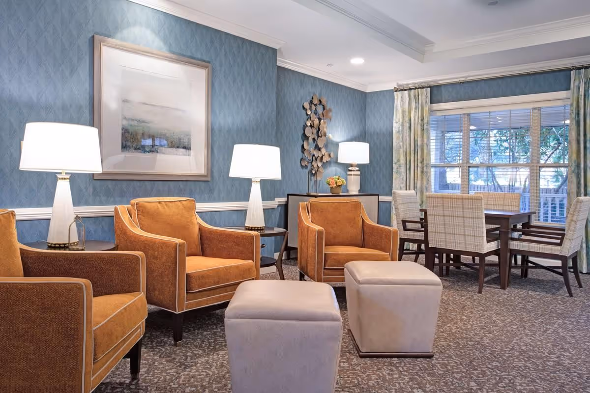 A cozy living room area with three orange armchairs, two beige ottomans, two side tables each with a white lamp, a framed abstract painting on a blue patterned wall, a decorative wall sculpture, and a dining table with six plaid upholstered chairs near a large window with floral curtains.