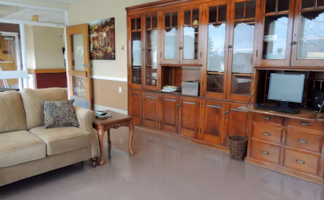 A cozy interior room featuring a beige sofa with a patterned cushion, a wooden side table with a book and remote control, and a large wooden cabinet with glass doors. A computer monitor and keyboard are placed on the right side of the cabinet. The room has a polished floor and a painting on the wall near an open door.