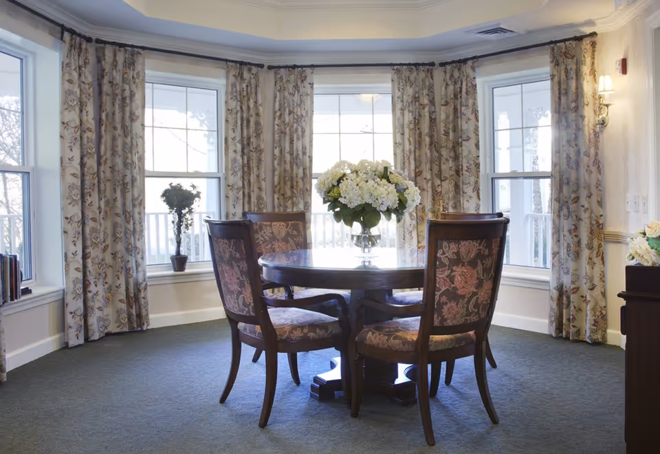 A bright dining area with a round wooden table surrounded by four upholstered chairs with floral patterns. The room features large windows with floral curtains, allowing natural light to fill the space. A vase with white flowers is placed in the center of the table, and there are small potted plants on the window sills.