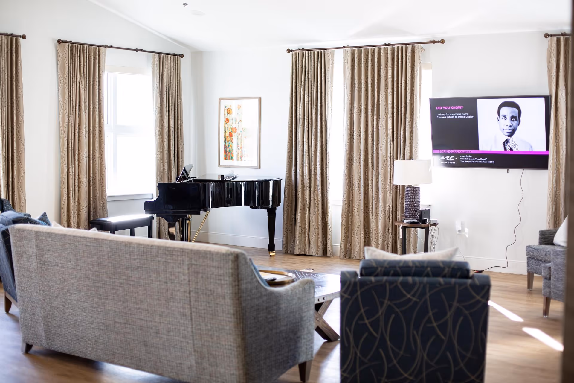 A bright living room with beige curtains covering windows, a black grand piano in the corner, a wall-mounted flat screen TV displaying a black and white photo of a man with text, a table lamp on a small side table, and several upholstered chairs and sofas arranged around a wooden coffee table on a wooden floor.