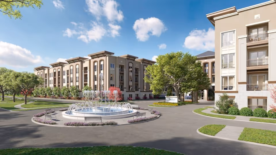 Exterior view of a senior living complex with a central circular fountain, landscaped courtyard, and multi-story buildings under a blue sky.