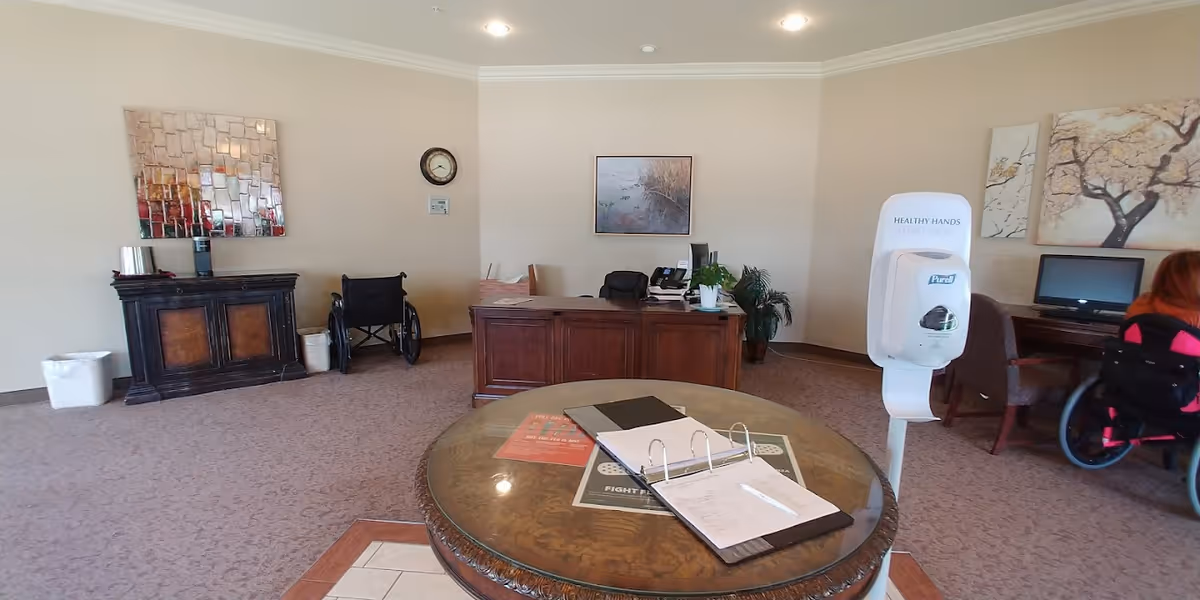 Reception area of a nursing and rehabilitation center with a wooden desk, a round table with papers and a binder, a wheelchair, a hand sanitizer dispenser, a computer station, and wall decorations including a clock and paintings.