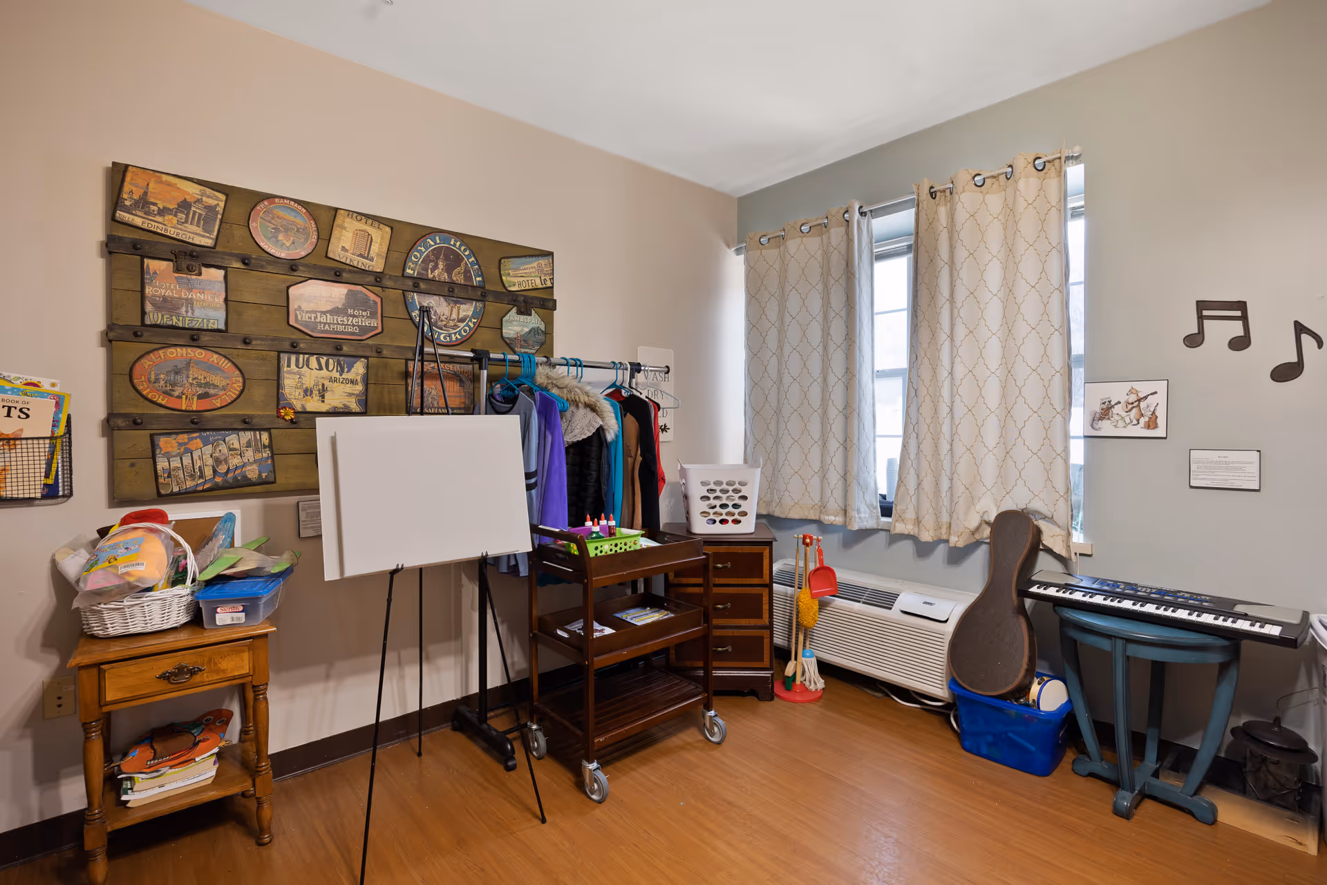 A cozy room with wooden flooring featuring a small wooden side table with baskets and books, a blank whiteboard on an easel, a clothing rack with coats, a rolling cart with art supplies, two windows with patterned curtains, a keyboard on a stand, a guitar case, and musical note wall decorations.