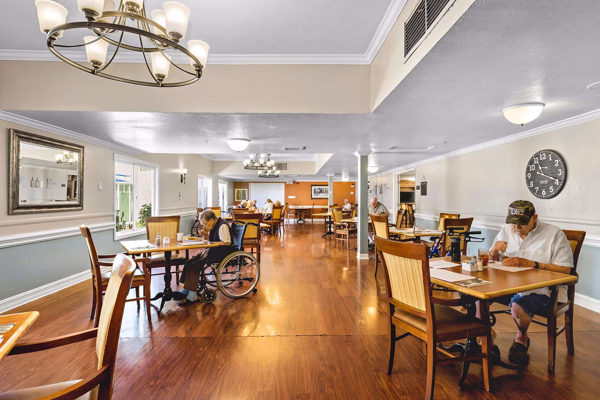 A spacious dining room in Montecito Assisted Living with wooden floors and multiple tables and chairs. Several elderly residents are seated at the tables, some eating and others engaged in quiet activities. The room is well-lit with ceiling lights and natural light from windows on the left side. The walls are painted in neutral tones with a large clock on the right wall.