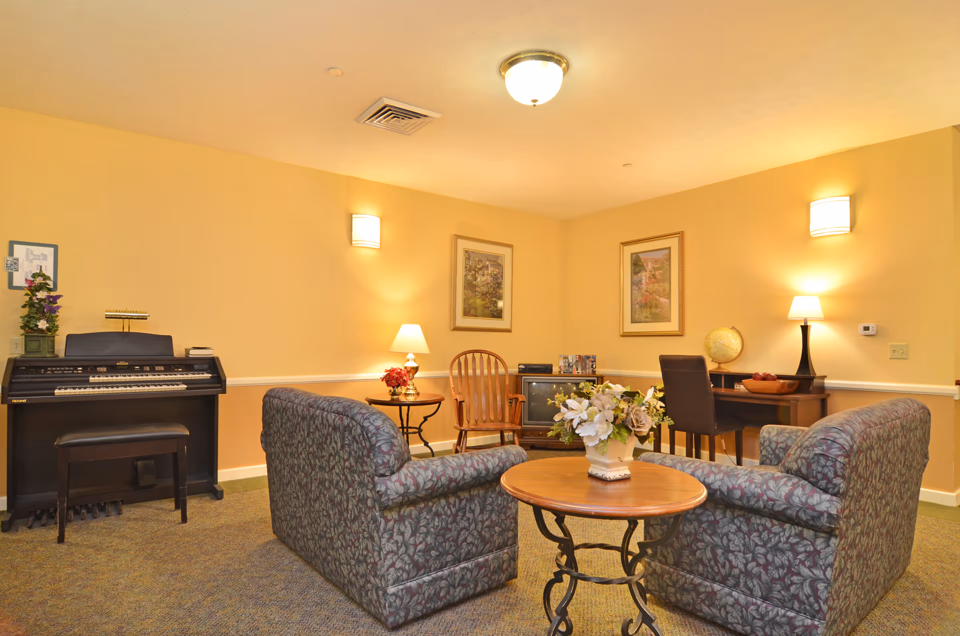 A cozy living room area with two patterned armchairs facing a round wooden table with a floral arrangement. The room has yellow walls with two framed pictures, a wooden rocking chair, a piano with a bench, a small TV on a stand, and a desk with a chair and a globe. The room is softly lit with wall sconces and table lamps.