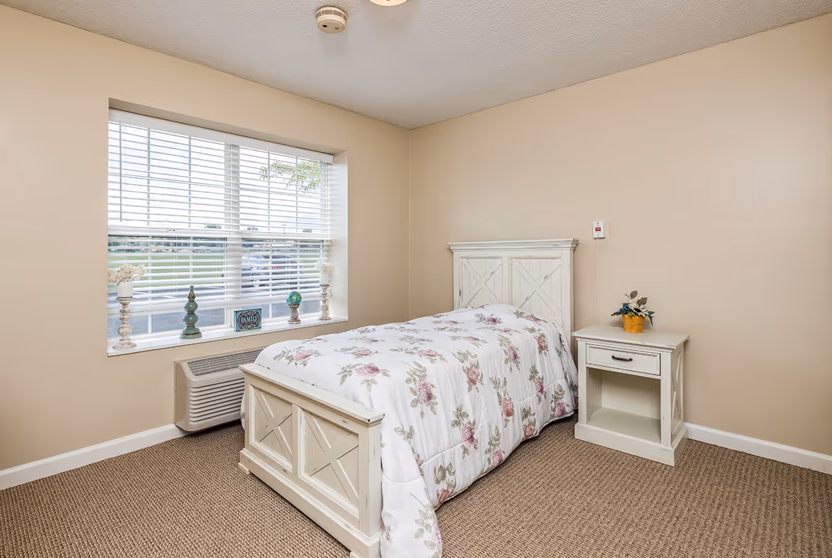 A small bedroom with beige walls and carpeted floor featuring a single bed with a white wooden frame and floral bedspread. Next to the bed is a matching white nightstand with a small potted plant on top. A large window with white blinds lets in natural light, and decorative items are placed on the window sill.