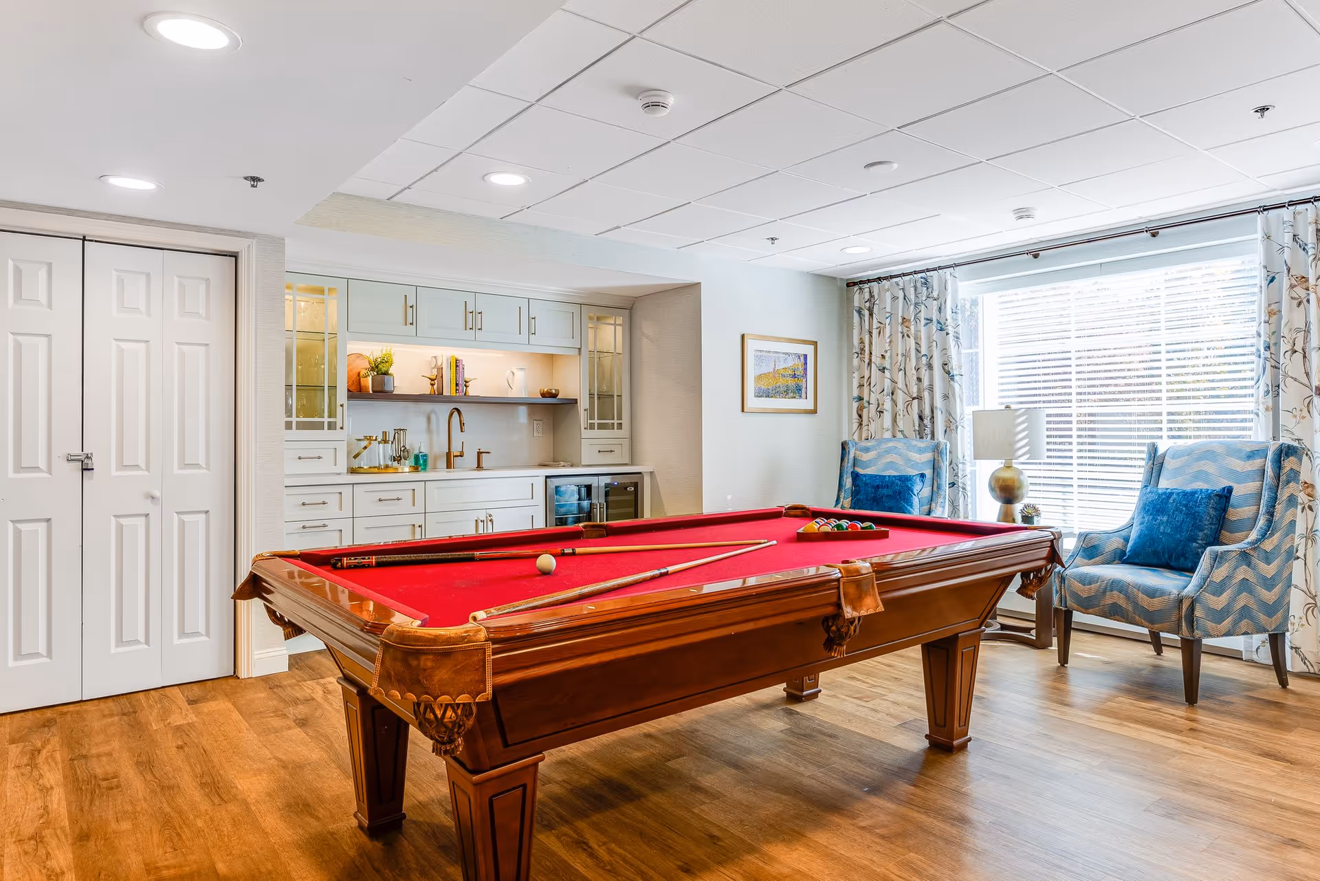 A bright recreational room featuring a red felt pool table with cues and balls arranged on it. Behind the pool table is a built-in cabinet with glass doors, shelves with decorative items, a sink, and a small wine cooler. Two blue patterned armchairs with blue cushions are placed near a large window with floral curtains, allowing natural light to fill the room. The floor is wooden, and the ceiling has recessed lighting.