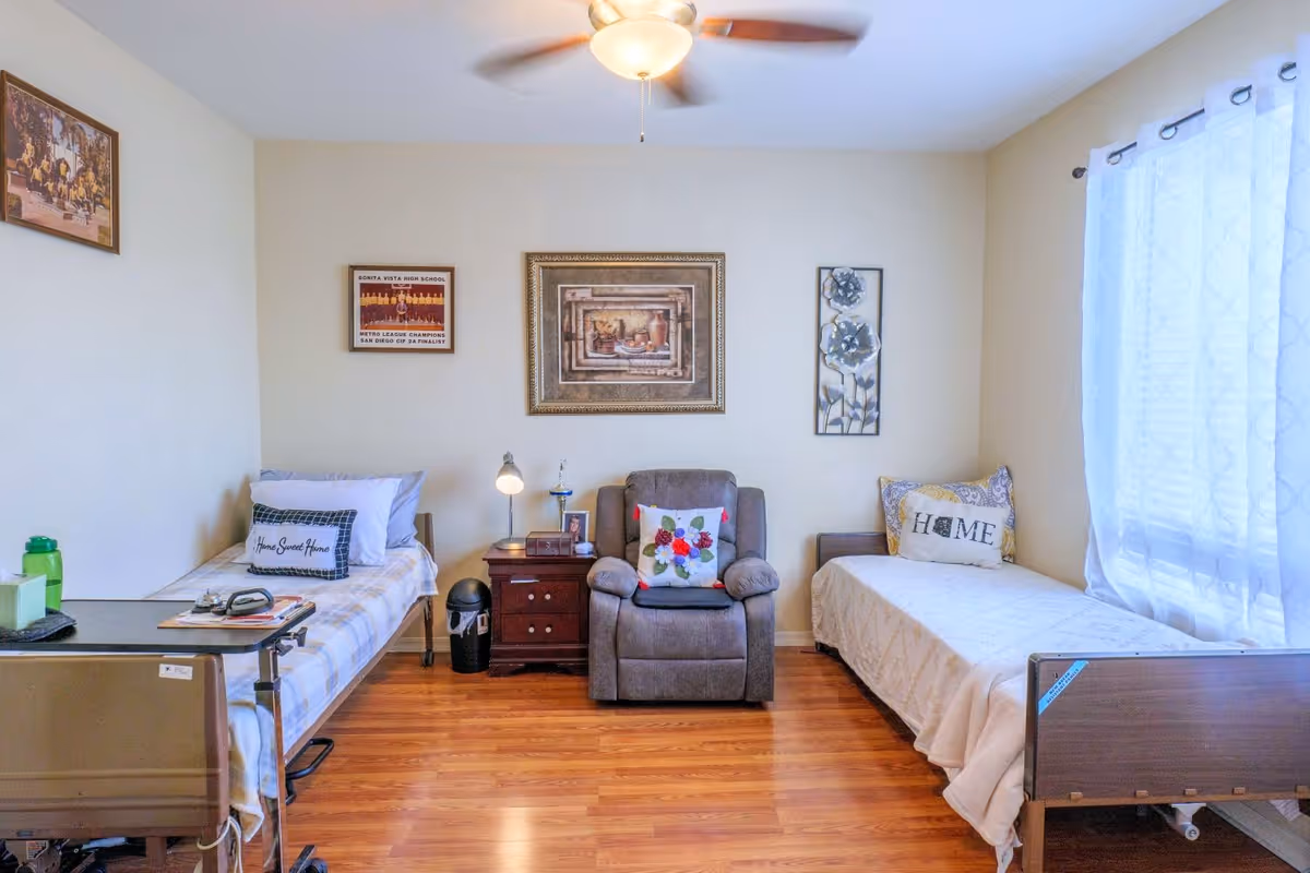 A bright and cozy room with two single beds on either side, each with pillows and blankets. Between the beds is a wooden nightstand with a lamp and some small items. A comfortable armchair with a floral cushion is placed in the center against the wall. The walls are decorated with framed pictures and artwork. A large window with sheer white curtains allows natural light to fill the room. The floor is wooden, and a ceiling fan with a light fixture is visible above.