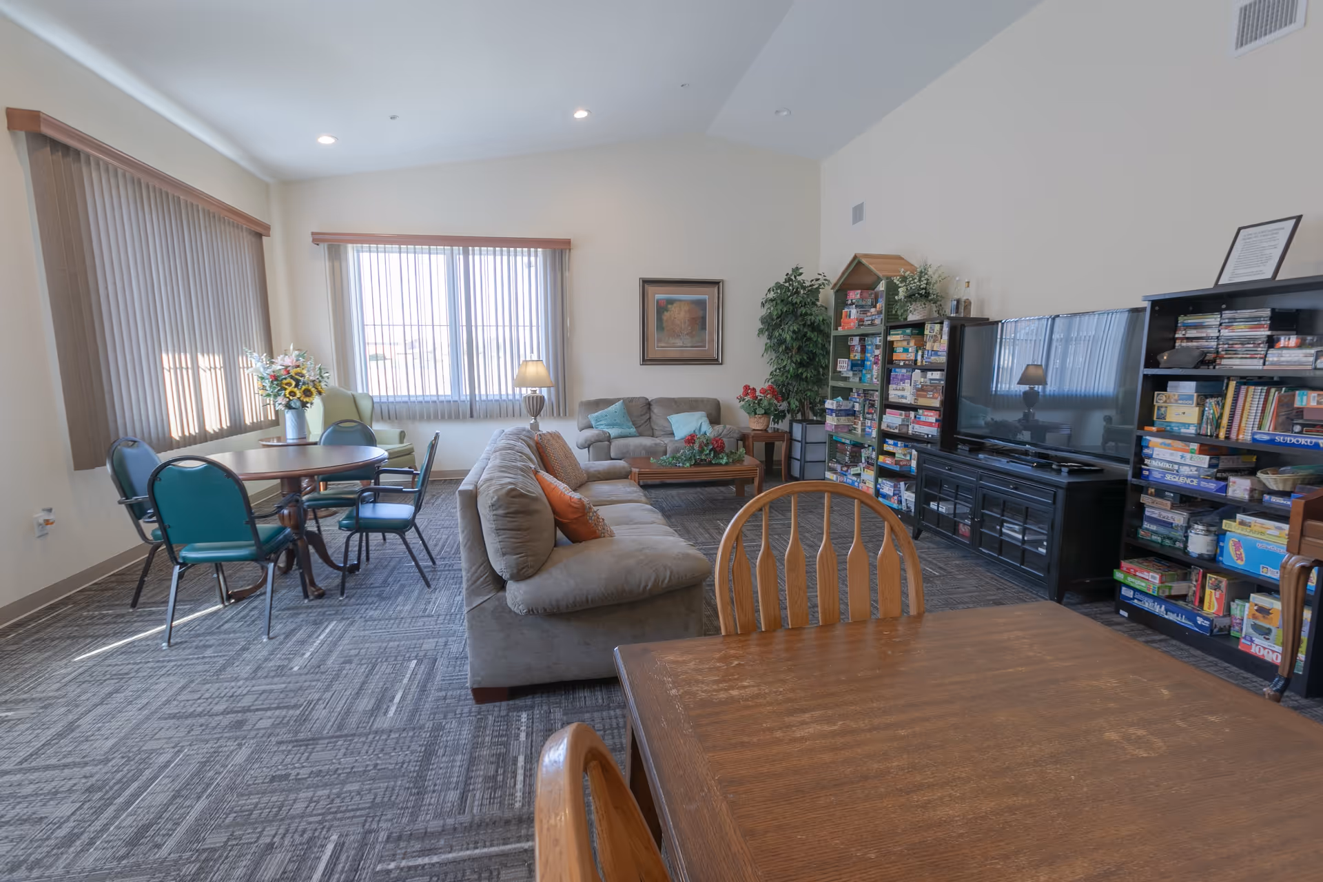 A cozy common area with a large beige sofa, a loveseat, and several chairs around wooden tables. There is a TV on a black stand with shelves filled with board games and books. The room has large windows with vertical blinds, a floor lamp, and decorative plants and flowers.