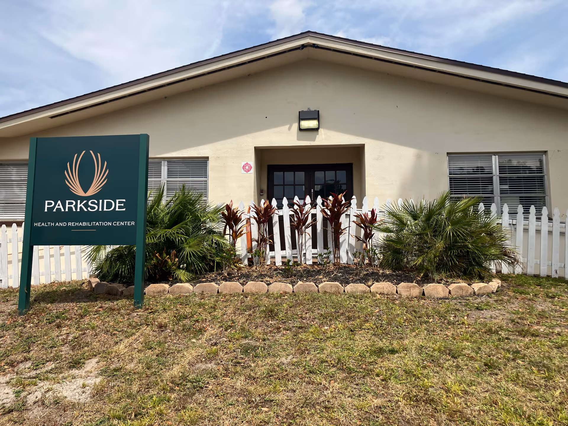Front exterior of Parkside Health and Rehabilitation Center with a green sign, white picket fence, and landscaped entrance.
