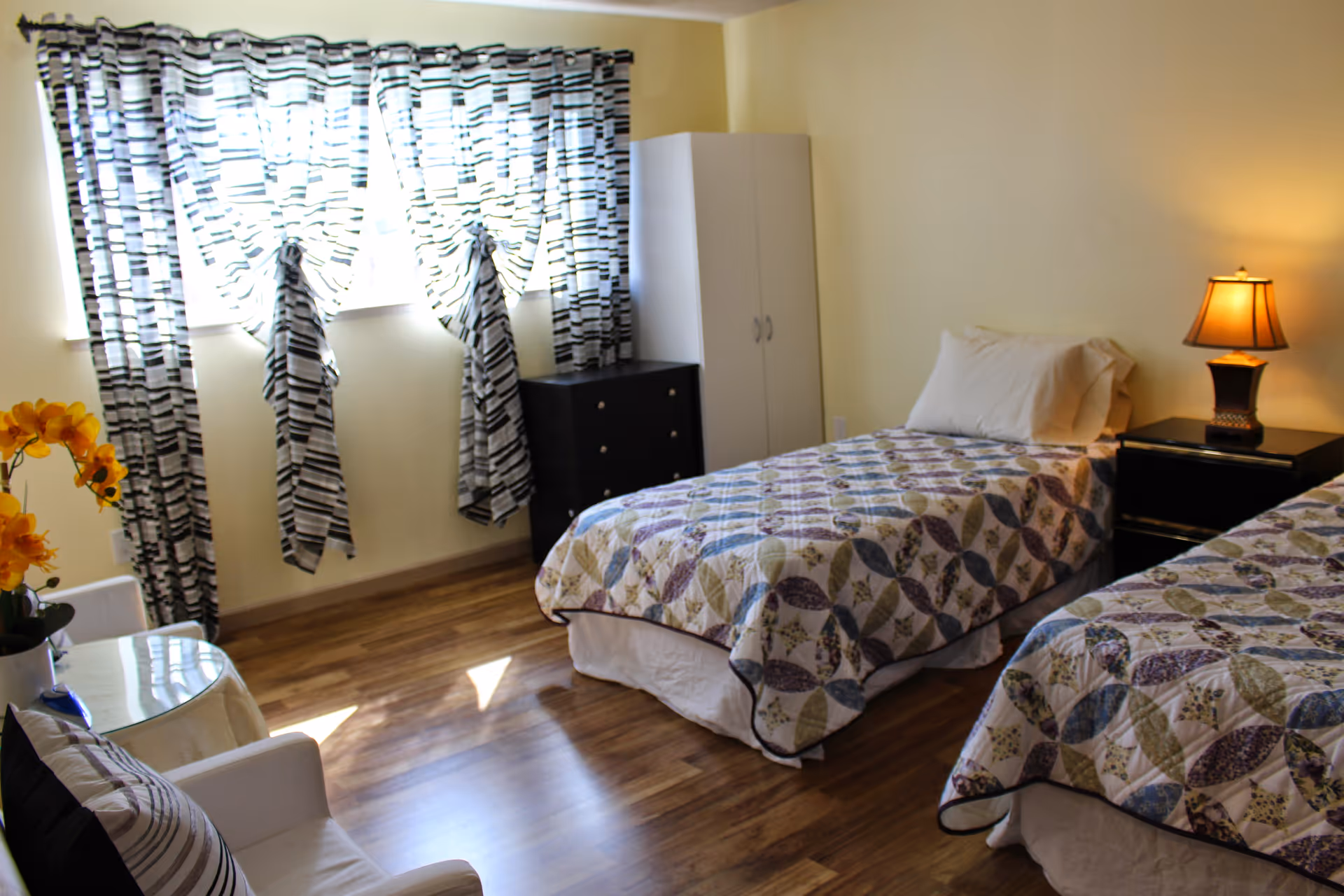 A bright bedroom with two single beds covered in patterned quilts, a nightstand with a lamp between the beds, a white wardrobe, a black chest of drawers, a white armchair with a striped pillow, and a window with black and white patterned curtains letting in natural light.