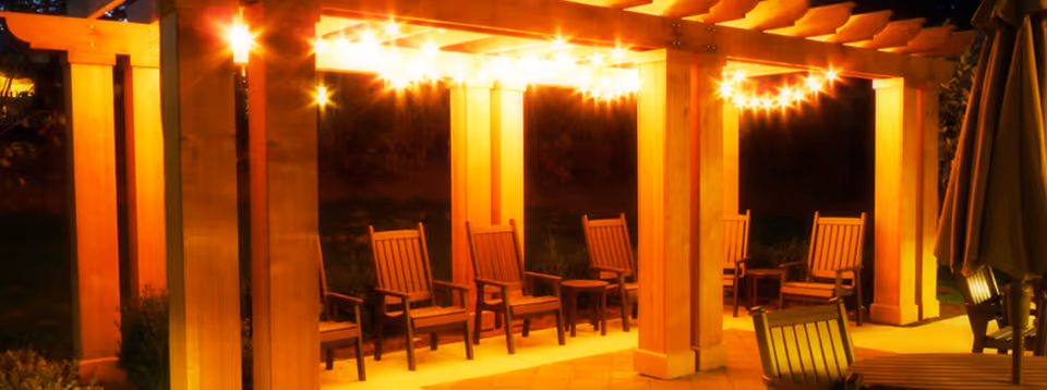 Wooden pergola over a patio illuminated by string lights with several wooden chairs and small tables at night.
